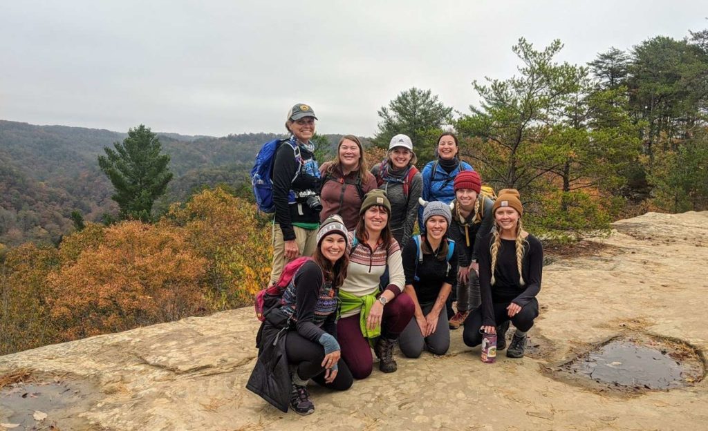 A group of hikers is posing on a rock ledge with a background of autumn-colored trees in a forested area