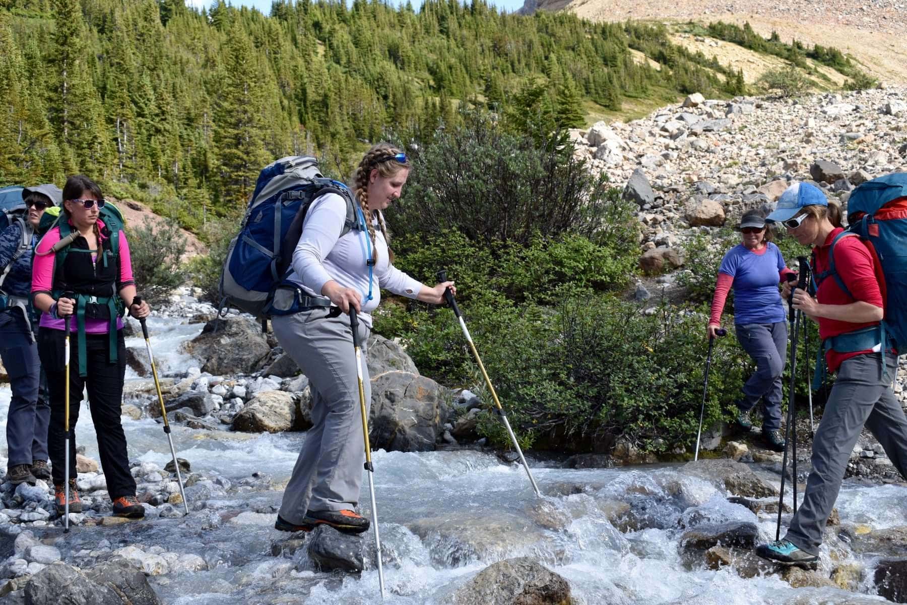 women walking on river rocks with the help of hiking poles