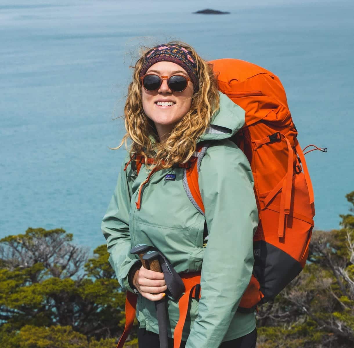 Courtney Stephenson hiking the W trek in Patagonia. She's smiling at the camera wearing an orange backpacking pack and holding trekking poles