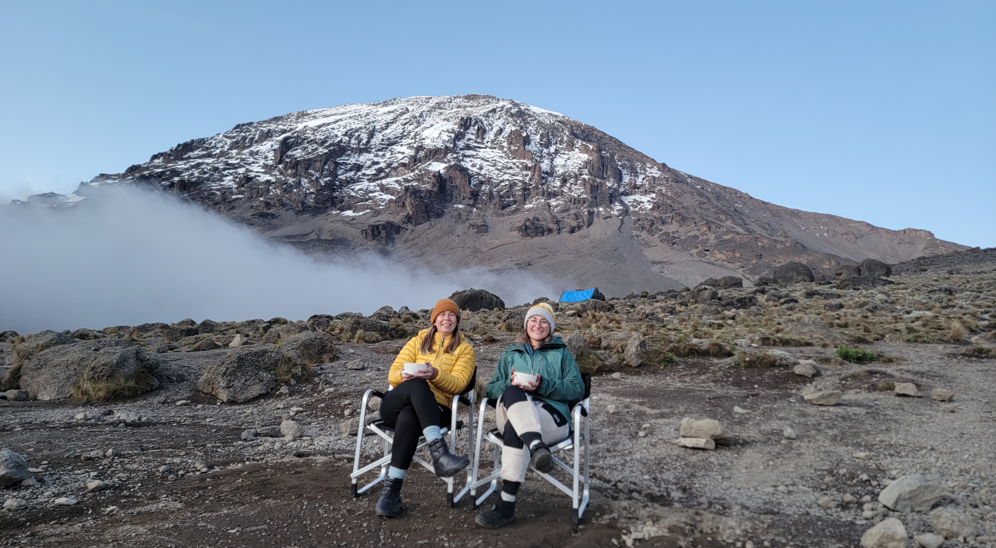 Two women enjoying hot drinks in camp chairs with snow-capped Mount Kilimanjaro rising behind them during Explorer Chick’s women’s climb.