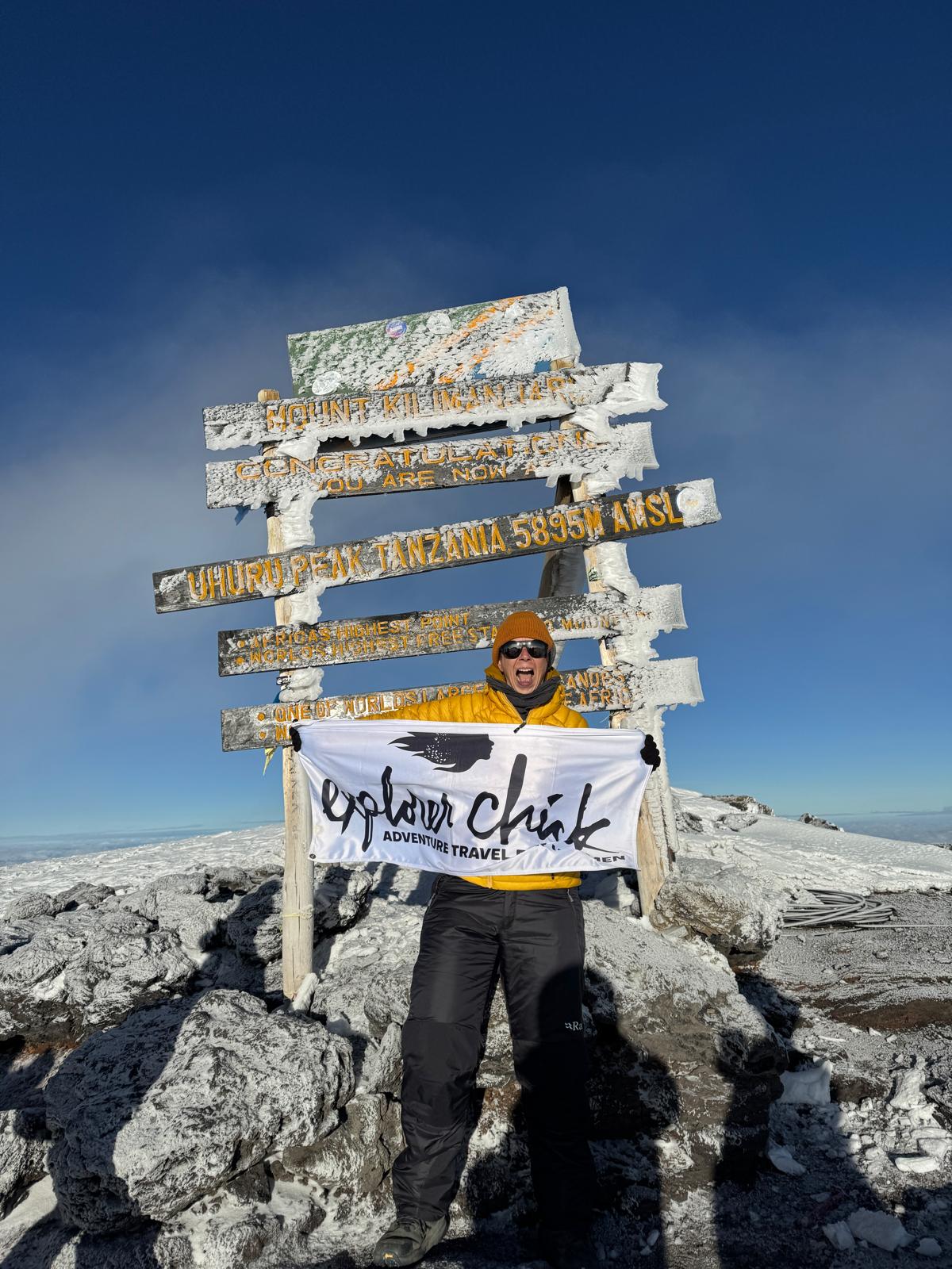 Woman holding Explorer Chick Adventure Travel flag at Uhuru Peak, the highest point of Mount Kilimanjaro, with snow and summit sign in background.