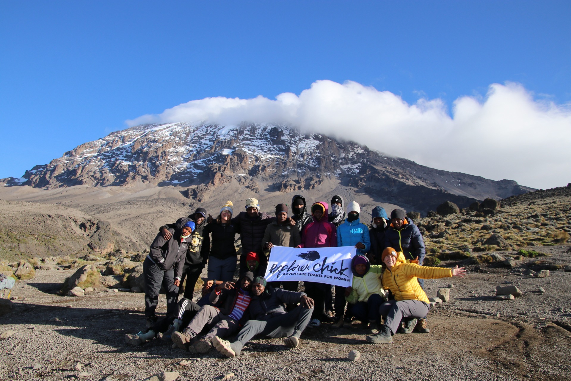 Group of women climbers and local guides holding an Explorer Chick banner with Kilimanjaro’s snowy slopes in the background.