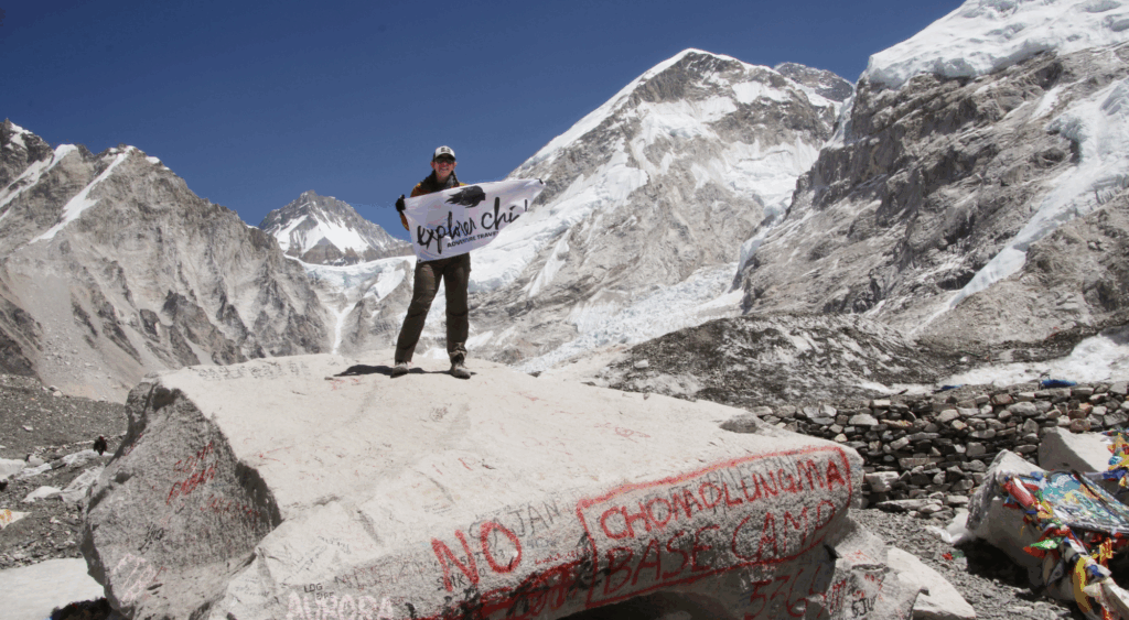 A woman holds an Explorer Chick flag on the marked Everest Base Camp rock with glaciers and Himalayan peaks in the background, celebrating the women’s group trekking adventure.