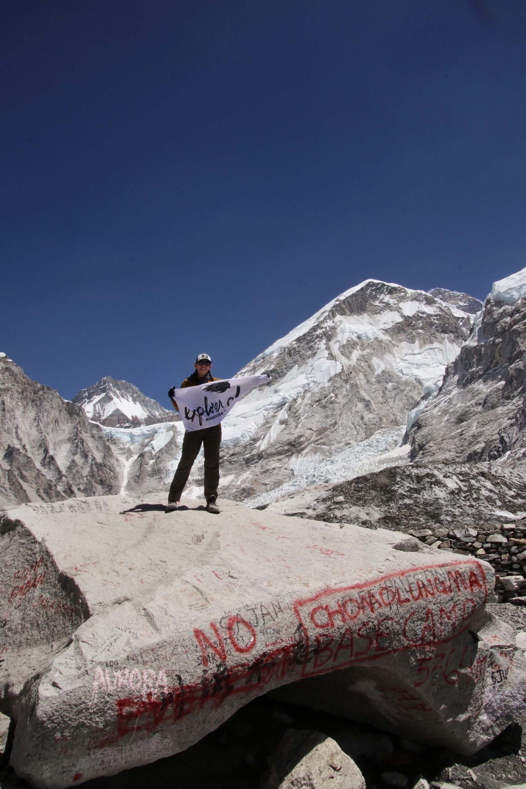 A female traveler stands on the iconic Everest Base Camp rock holding an Explorer Chick flag, with glaciers and snowcapped peaks rising behind her in Nepal.