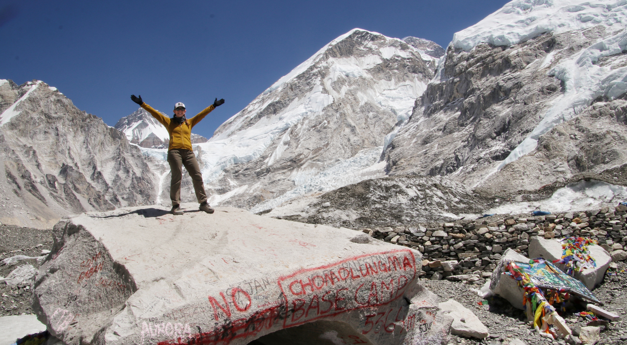 A smiling woman raises her arms on the iconic Everest Base Camp rock, surrounded by prayer flags, glaciers, and snowy Himalayan peaks.
