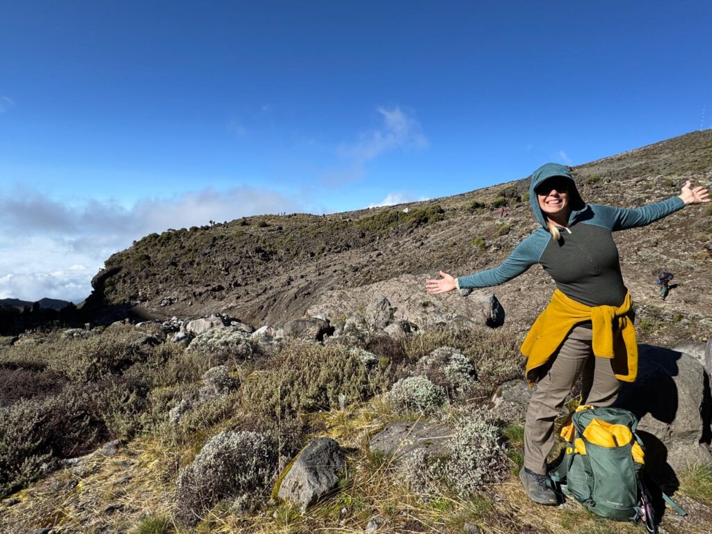 Woman hiker posing with arms wide open on the Kilimanjaro trail with alpine landscape and blue skies in the background.