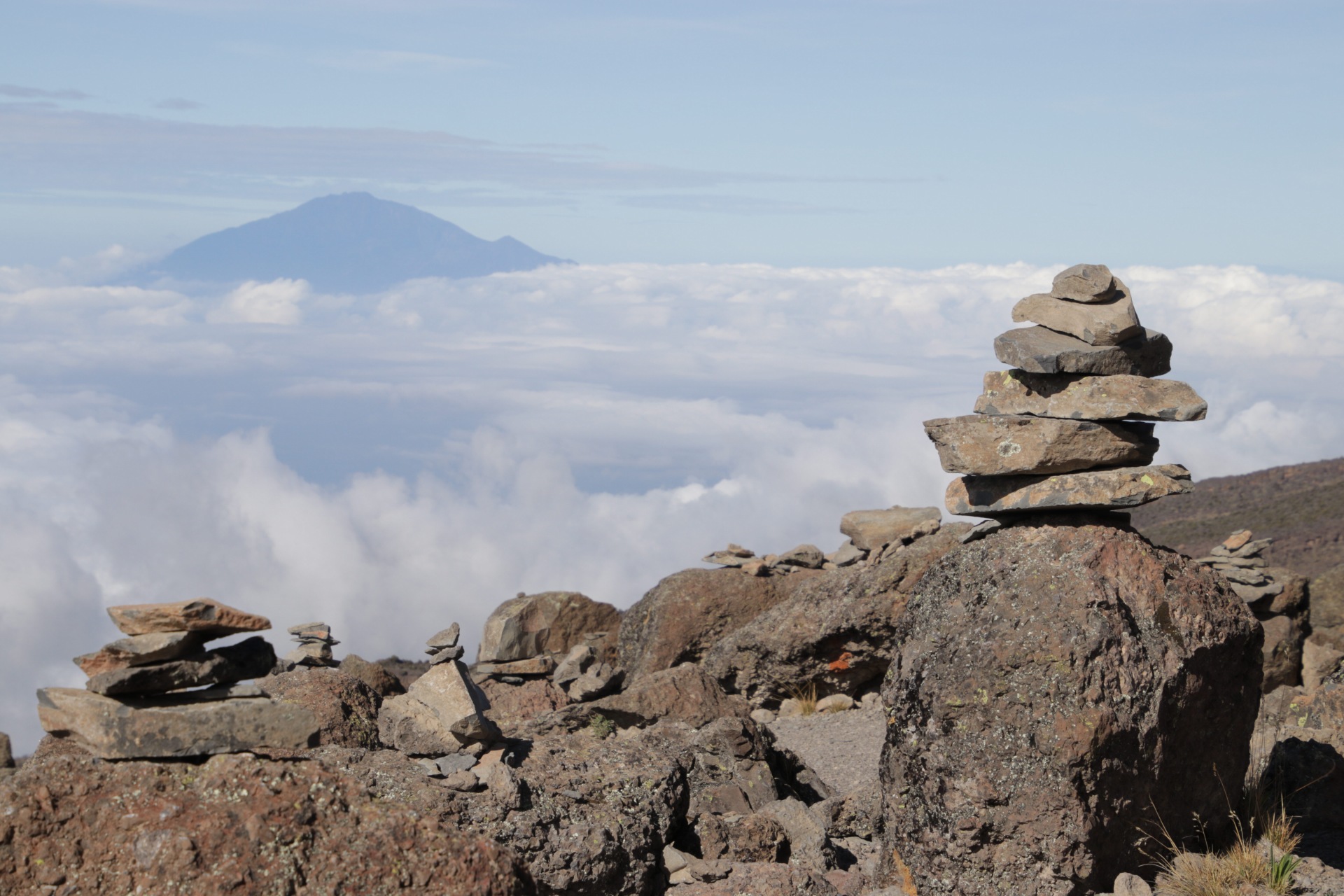 Stone cairns stacked along the Kilimanjaro trail with mountain views above the clouds in the distance.