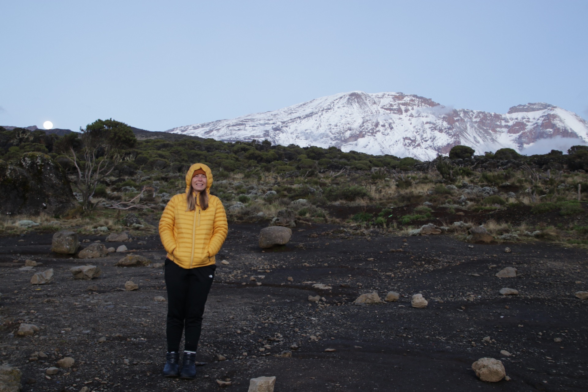 Woman in yellow puffy jacket standing in front of snow-covered Kilimanjaro summit during Explorer Chick’s women’s climb.