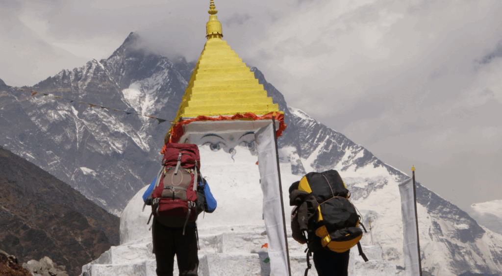 Two trekkers with large backpacks walk past a white Buddhist stupa with painted eyes and a golden spire, set against the backdrop of the Himalayas.
