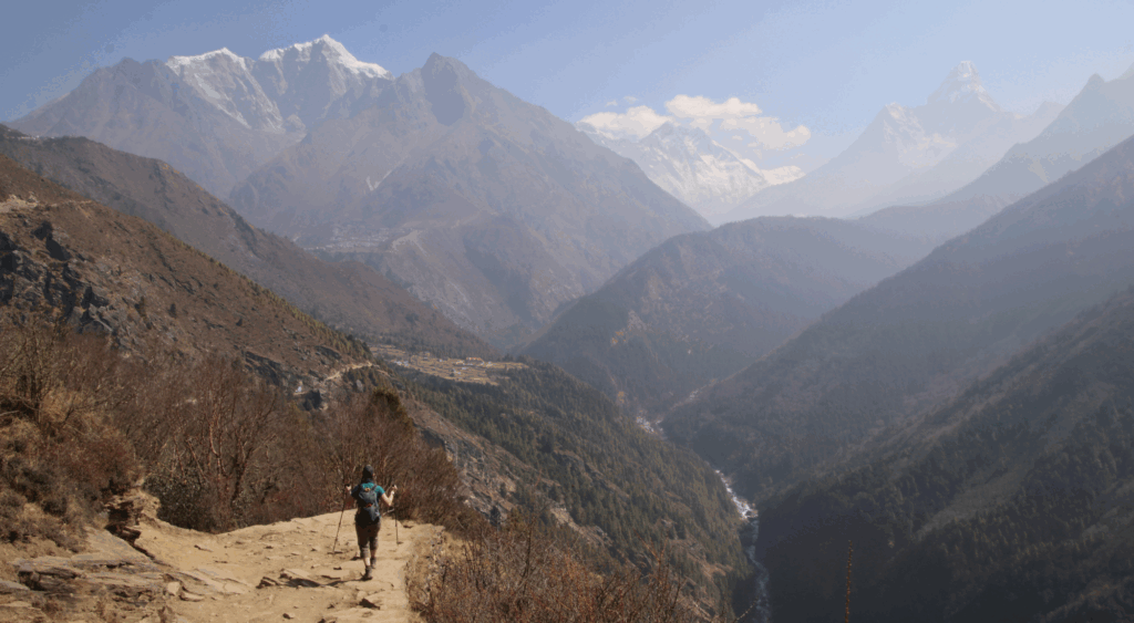 A solo hiker with trekking poles walks along a dirt trail toward Everest Base Camp, surrounded by deep valleys and snowcapped peaks of the Himalayas.