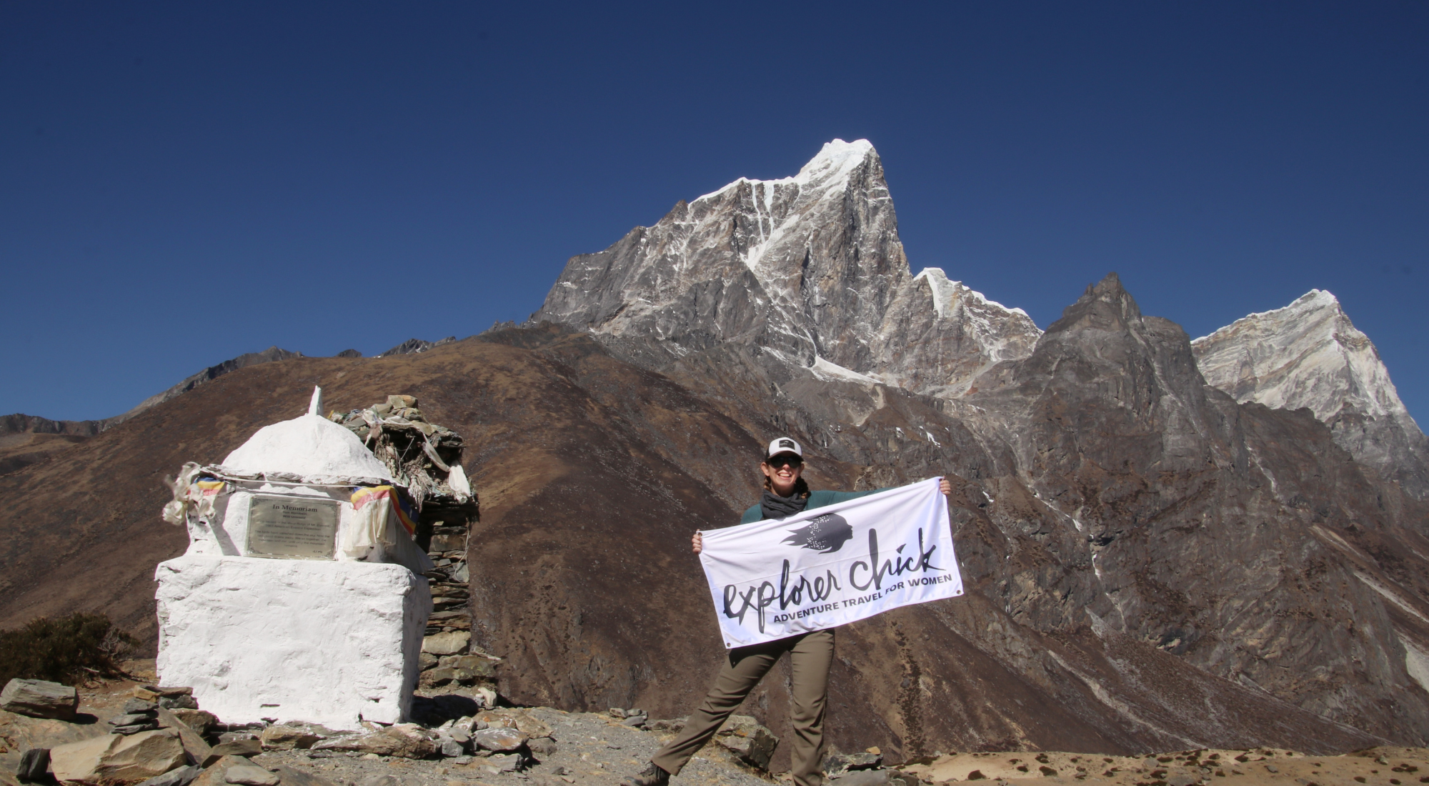 A smiling woman holds an Explorer Chick flag beside a white stupa, with jagged Himalayan peaks rising behind her on the Everest Base Camp trail.
