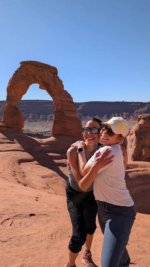 Two women smiling and hugging in front of Delicate Arch in Arches National Park, Utah, during an Explorer Chick women’s hiking adventure under a bright blue sky.