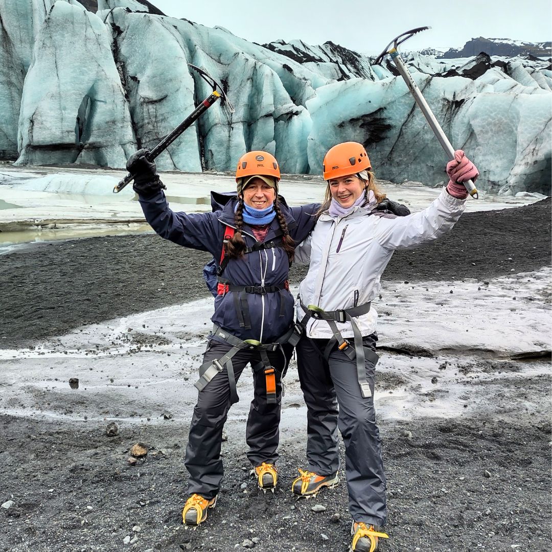 Two women glacier hiking in Iceland wearing helmets and crampons, posing with ice axes in front of a blue ice glacier during an adventurous outdoor excursion