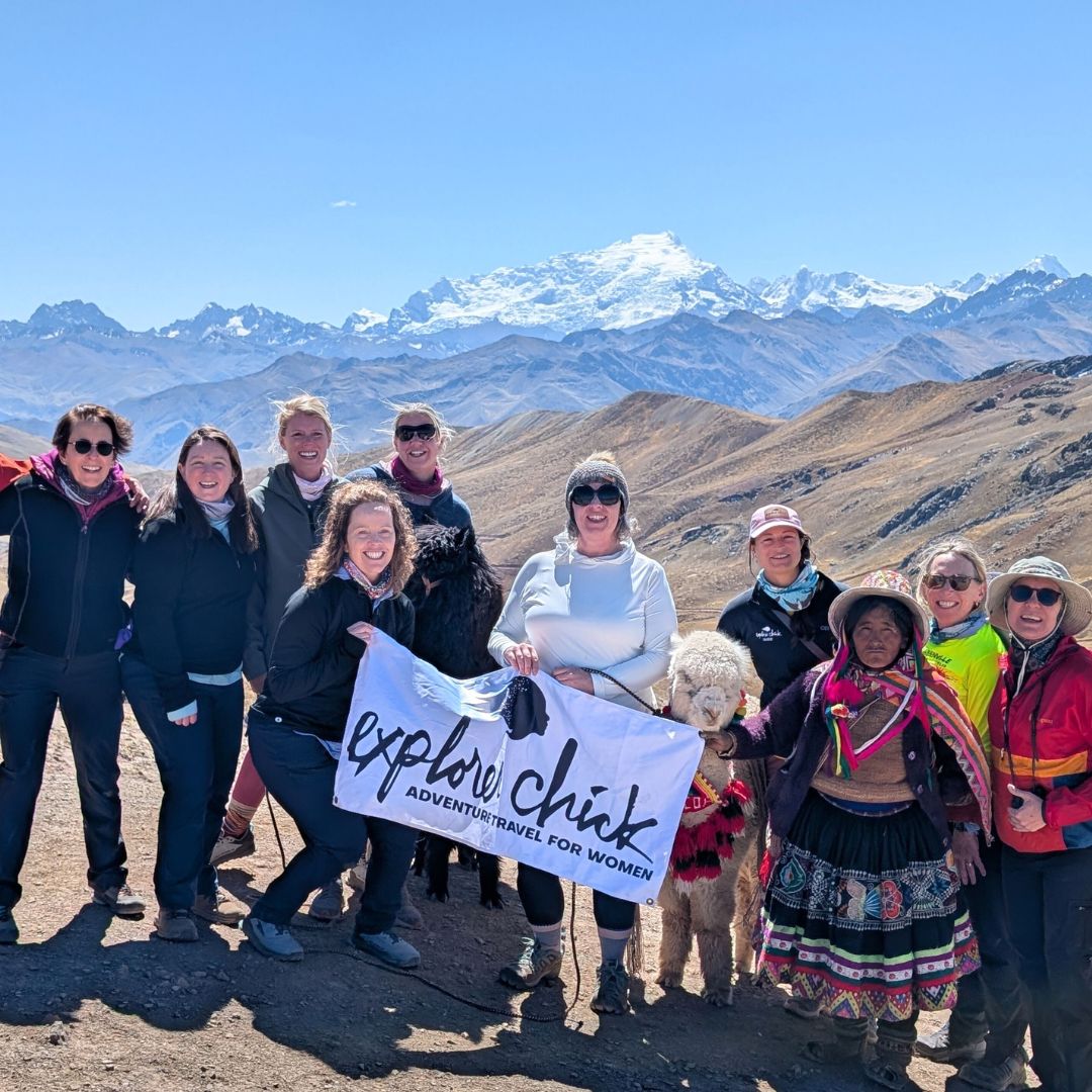Explorer Chick women’s hiking group posing with local guides and llamas in the Peruvian Andes with snow-capped mountains in the background